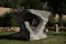 A large, abstract stone sculpture situated outdoors on a grassy area. The sculpture features a unique design with a hollow center and curved, textured surfaces. It is surrounded by greenery, including trees and bushes, and there is a stone wall in the background. The setting appears to be a garden or park, providing a natural backdrop to the artistic piece.