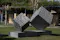 The image shows a public art installation featuring two large, intricately designed cubes with a maze-like pattern on their surfaces. These cubes are positioned on a pedestal in an outdoor setting, surrounded by grass and trees, suggesting a park or garden environment. The artwork appears to be a modern sculpture, emphasizing geometric shapes and complex patterns.