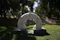 A modern sculpture situated in a park-like setting. The sculpture is made of stone and features an abstract, circular design with smooth, flowing curves. It is placed on a pedestal and surrounded by grass and trees, suggesting a serene and natural environment. The artwork appears to be a contemporary piece, blending artistic expression with the tranquility of nature.