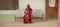 This image shows a woman wearing a red hijab and dress sitting on a metal bench outside a building, holding a young child on her lap. The child has small braids with colorful beads and is wearing a blue and white striped outfit. The woman looks off to the side with a slightly smiling expression, while the child gazes downward. The setting appears to be a waiting area outside a clinic or medical facility, as indicated by a poster on the door in the background. The poster features an illustration of a doctor and text encouraging people with cough symptoms to seek free treatment. The walls of the building are painted in a faded green color, and the door is slightly damaged at the bottom. The ground is tiled, and the area is well-lit, suggesting it is daytime.