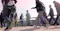 The image shows a group of people walking outdoors on a sunny day. They are dressed in a mix of casual and business attire, suggesting a busy urban environment. The scene captures the movement and diversity of individuals in a public space.