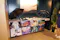 A display table with Tokyo-themed posters, umbrellas, and various promotional items is set up at an event. Posters feature traditional and modern Tokyo imagery, including Kabuki, robots, historic sites, and anime. A woman stands to the right of the table.
