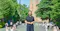 The image shows her standing outdoors in front of a prominent clock tower, surrounded by students and greenery.