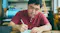 An artist, dressed in a red shirt, is focused on drawing at a desk surrounded by art supplies, illustrating deep engagement in the creative process.