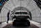 The image shows a TOGG electric vehicle undergoing inspection or quality control in a modern, well-lit automotive facility. The car is positioned in a tunnel with bright, curved LED lights, highlighting its sleek design and advanced features. Two workers are examining the vehicle, indicating a focus on precision and quality in the production process. The overall setting emphasizes innovation and high standards in automotive manufacturing.