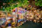 The image shows a vibrant indoor floating market scene, likely located within a shopping mall or themed attraction in Thailand. The setting features traditional wooden boats on a waterway, decorated columns, colorful lanterns hanging from the ceiling, and market stalls selling various goods. The atmosphere is lively and festive, recreating the experience of a Thai floating market with a blend of modern and traditional architectural elements. This immersive environment is designed to showcase Thai culture and provide visitors with a unique shopping and dining experience.