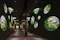 A dark corridor with circular and oval windows in the walls, each revealing views of green leafy trees outside. The reflections and soft lighting evoke a peaceful, immersive forest-like experience.