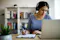 The image shows a person sitting at a desk, wearing headphones, and writing on a clipboard while looking at a laptop. In the background, there is a bookshelf filled with books, and a potted plant is visible in the foreground. The setting appears to be a home or office environment, suggesting the person is engaged in studying, remote work, or an online meeting.