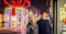This image shows two people standing on a city street at night during the winter season. They are dressed warmly in jackets and hats, and appear to be looking at a shop window display. In the background, there is a large, illuminated holiday decoration shaped like a gift box with a bow, surrounded by festive lights, suggesting a holiday or Christmas atmosphere. The street is wet, possibly from recent rain or snow, and the overall scene conveys a cozy, festive mood.