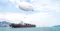 This image shows a large cargo ship on the ocean, loaded with shipping containers. Above the ship, a modern airship or blimp labeled "FLYING WHALES" is flying, carrying two shipping containers suspended beneath it. The scene suggests innovative logistics and transportation methods, possibly highlighting the use of airships for heavy-lift cargo transport over water or to remote areas. In the background, there are mountains, city buildings, and other ships, indicating a busy port or coastal region.