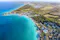 An aerial view of a coastal landscape shows turquoise waters, sandy beaches, and a sprawling resort complex surrounded by lush greenery and fields. The coastline curves gently, with distant hills visible on the horizon.