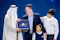 A man in a dark suit receives an award from a man in traditional Middle Eastern attire. Two children stand beside them, looking happy. The mood is celebratory.