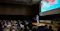 A speaker stands on a stage in a conference hall, addressing an audience. The audience is seated and attentively listening. A large screen behind the speaker displays information about an upcoming congress in Tokyo, with text in both English and Japanese. The hall is modern, with high ceilings and professional lighting.