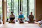 Three people are seated cross-legged on cushions in a traditional Japanese room with tatami mats. They are positioned in front of large glass doors that open to a lush garden outside. Their hands rest in their laps in a meditative posture. The room is serene and filled with natural light from the garden.