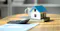 The context is real estate or home finance. The image shows a small model house with a blue roof placed on top of a notebook, alongside a calculator and some paperwork on a wooden table. This setting suggests discussions or activities related to buying, selling, or financing a home, such as mortgage planning, property investment, or real estate management.