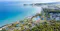 A large expanse of turquoise blue sea on the left, with the coastline curving gently. Along the coast, there are sandy beaches and several resort buildings, many with green roofs and surrounded by lush greenery and trees. There are multiple swimming pools visible, as well as landscaped gardens and pathways connecting the various buildings. The resort area is bordered by dense forest, and the horizon stretches out with more land and sea in the distance. The overall scene is bright, serene, and inviting, with a mix of natural and built environments.