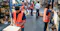 The image shows several workers in a warehouse environment. They are wearing orange safety vests and are engaged in tasks such as packing boxes, organizing items on shelves, and managing inventory. The warehouse is filled with shelves, boxes, and various supplies, indicating a busy and organized workspace focused on logistics or distribution activities.