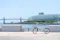 A white folding bicycle with a basket stands on a paved waterfront area, with a modern glass building, a bridge, and a ship in the background.