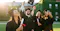 A group of graduates is celebrating outdoors, wearing caps and gowns. Some are holding diplomas, one is taking a selfie, and another is tossing their graduation cap into the air. The scene is set in front of a university building covered in ivy, with sunlight illuminating the moment, signifying achievement and joy.