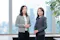 Two women in business attire stand side by side in a modern office with large windows and a city view. The woman on the left wears a patterned jacket and white top; the woman on the right wears a grey blazer and black dress. Both have their faces blurred for privacy.
