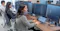 A group of people work at desks in a modern office, each using multiple computer monitors. The focus is on a woman in the foreground typing on a laptop connected to two large screens displaying code. Other colleagues are visible in the background, all seated in ergonomic chairs, with their faces blurred for privacy. The workspace is bright and professional, with large windows letting in natural light.