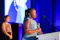 Woman holds a glass award while speaking at a podium during the .ORG Impact Awards, with blue stage lighting and another attendee blurred in the background.