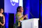Woman holding a glass award speaks at a podium during the .ORG Impact Awards, with blue stage lighting and another attendee standing blurred in the background.