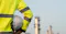 Worker in a high-visibility yellow safety jacket holding a white hard hat, standing in front of a blurred industrial oil refinery.