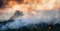 Helicopter dropping water over a smoky forest wildfire at sunset, with flames visible among trees