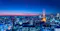 Panoramic night view of a city skyline with brightly lit skyscrapers and buildings. A tall, illuminated orange tower stands prominently on the right side, surrounded by city lights. The sky is a gradient of blue and orange hues, indicating dusk, and distant mountains are visible on the horizon.