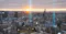 A cityscape at sunset with tall buildings and mountains in the background. Digital graphics overlay the image, representing a connected smart city with icons for people, networks, and technology, as well as glowing vertical lines indicating data or connectivity points throughout the city.
