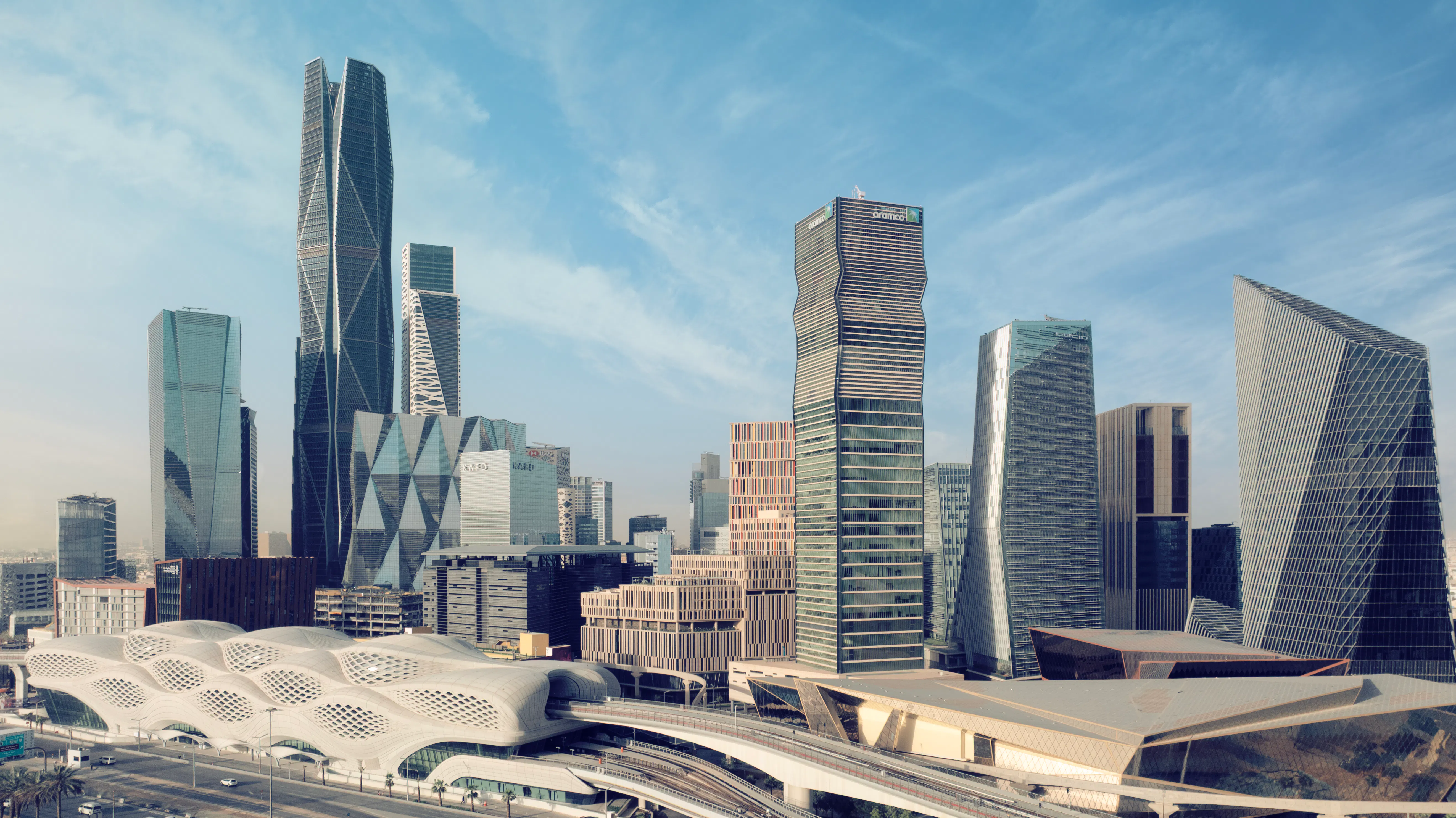 Panoramic view of a modern city skyline with futuristic glass skyscrapers, a white wave-shaped transit station in the foreground, and a blue sky streaked with thin clouds.
