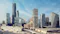Panoramic view of a modern city skyline with futuristic glass skyscrapers, a white wave-shaped transit station in the foreground, and a blue sky streaked with thin clouds.