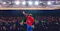 Speaker standing on a conference stage facing a large audience, raising one hand beneath bright overhead lights in a packed event venue, illustrating a keynote presentation, industry conference, or leadership event.