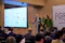 A man stands at a clear podium, speaking to an audience at the FinCity.Tokyo Global Forum. A large screen shows a list of member organizations. The event is held in a modern conference room with wooden walls and green plants.
