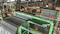 An industrial facility interior showing large green machinery labeled "CATHOLYTE MIDDLE." The machines have multiple metal plates and tubes connected to them, with various pipes and cables running throughout the area. The facility is spacious, with metal beams, windows, and other equipment visible in the background, indicating a complex manufacturing or processing operation.
