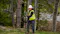 A utility worker wearing a white hard hat and high-visibility safety vest carries a coil of black cable across a grassy area beside a wooded roadside.