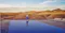 Person walking beside a reflective infinity pool overlooking rolling desert hills and distant mountains beneath a clear blue sky.