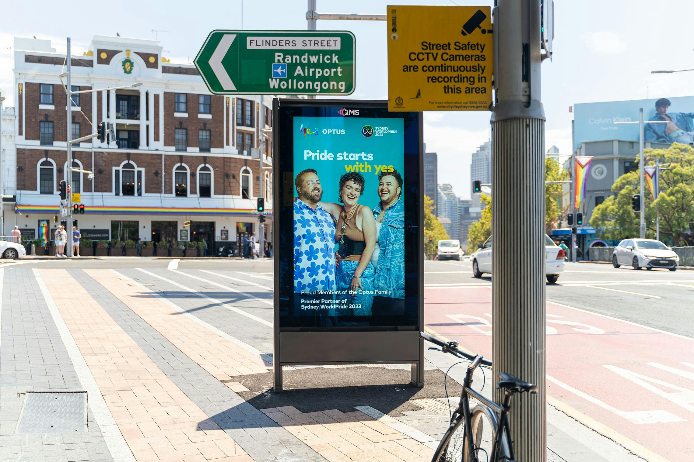 A photo of a street furniture placement on Taylor Square featuring the Optus x SydneyWorldPride campaign: Pride starts with yes