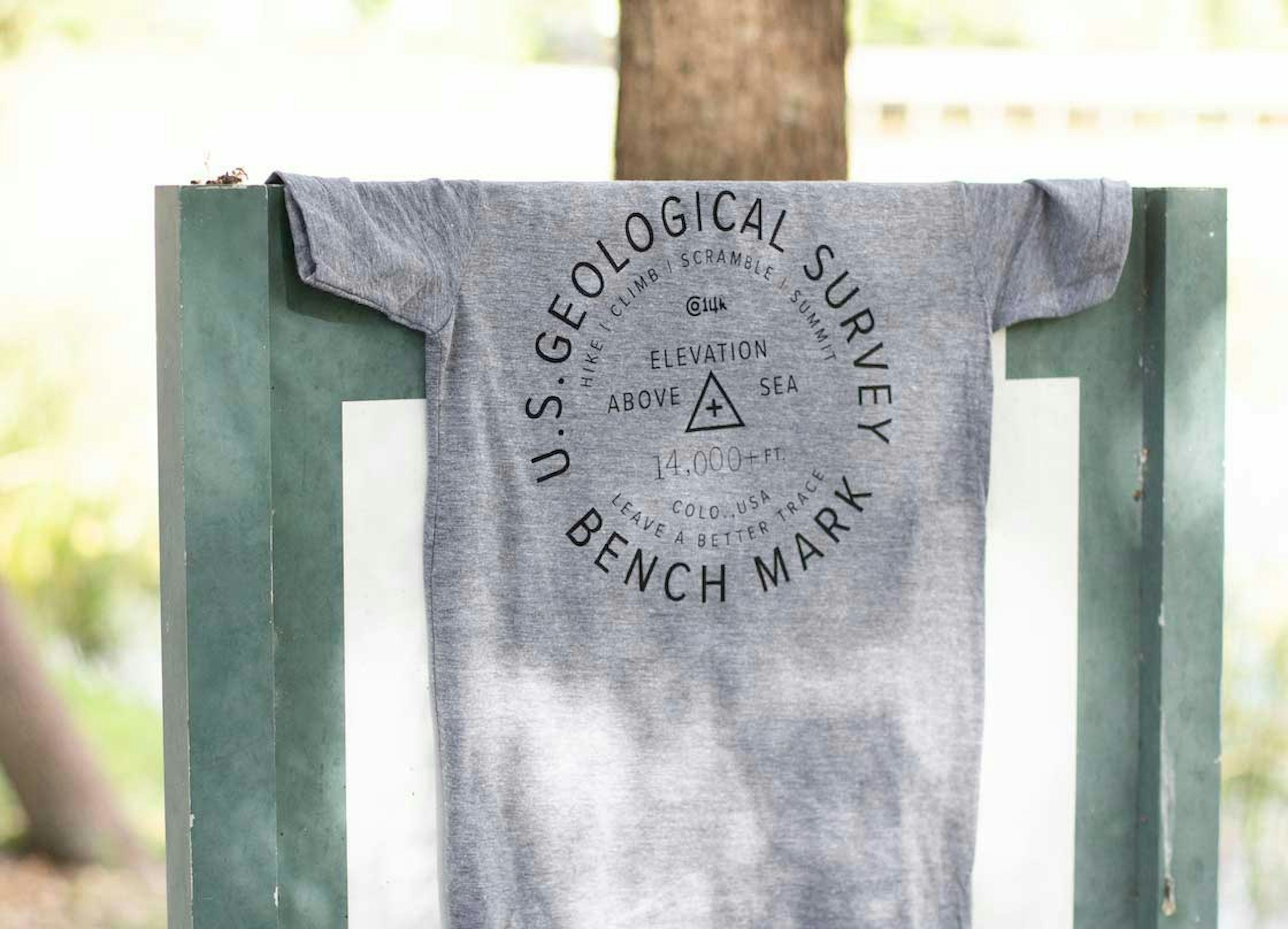 A gray shirt that says US Geological Survey Benchmark rest atop a bench in a park