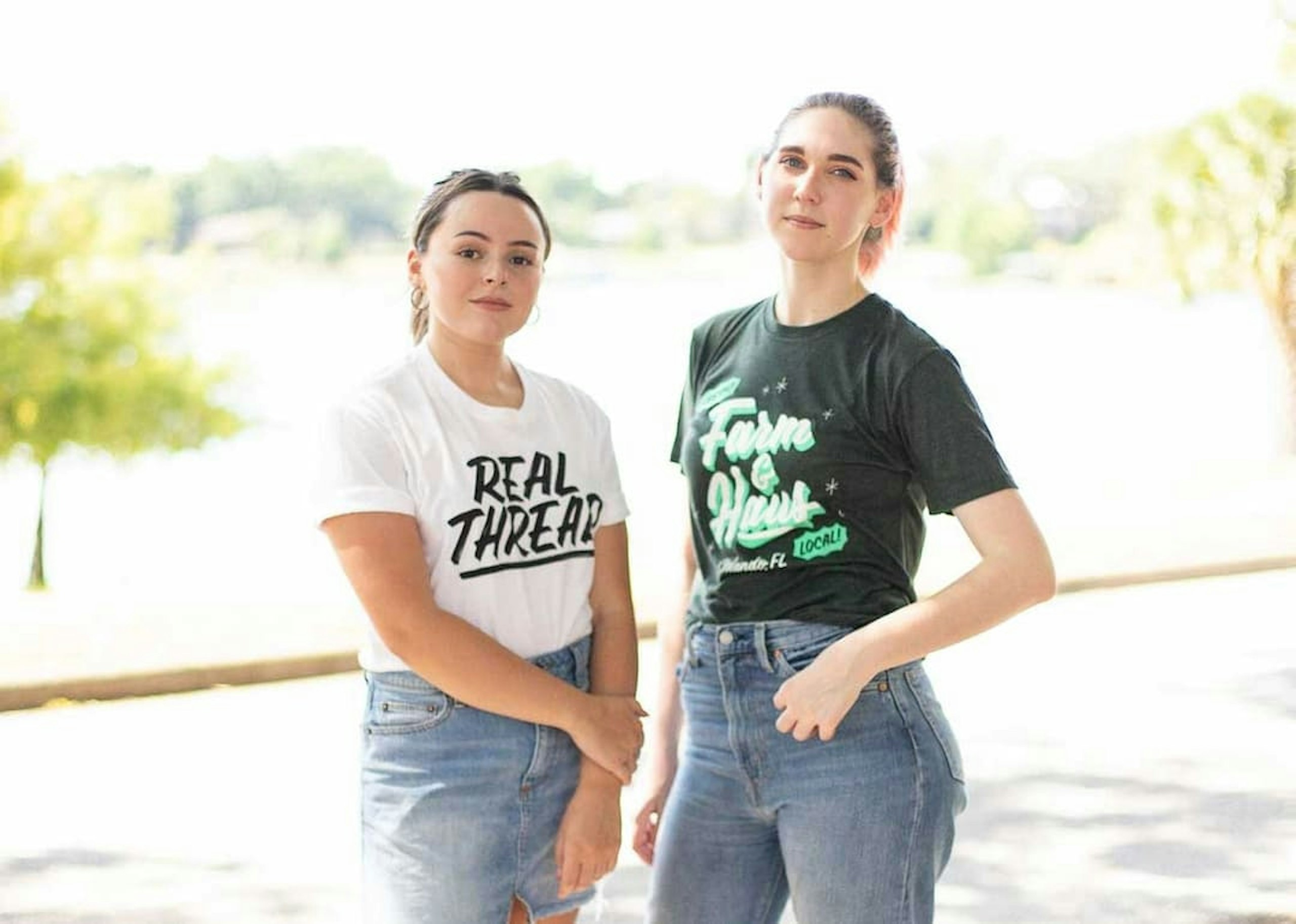One woman wearing a white shirt with Real Thread on it and one woman wearing Farm and Haus Shirt.