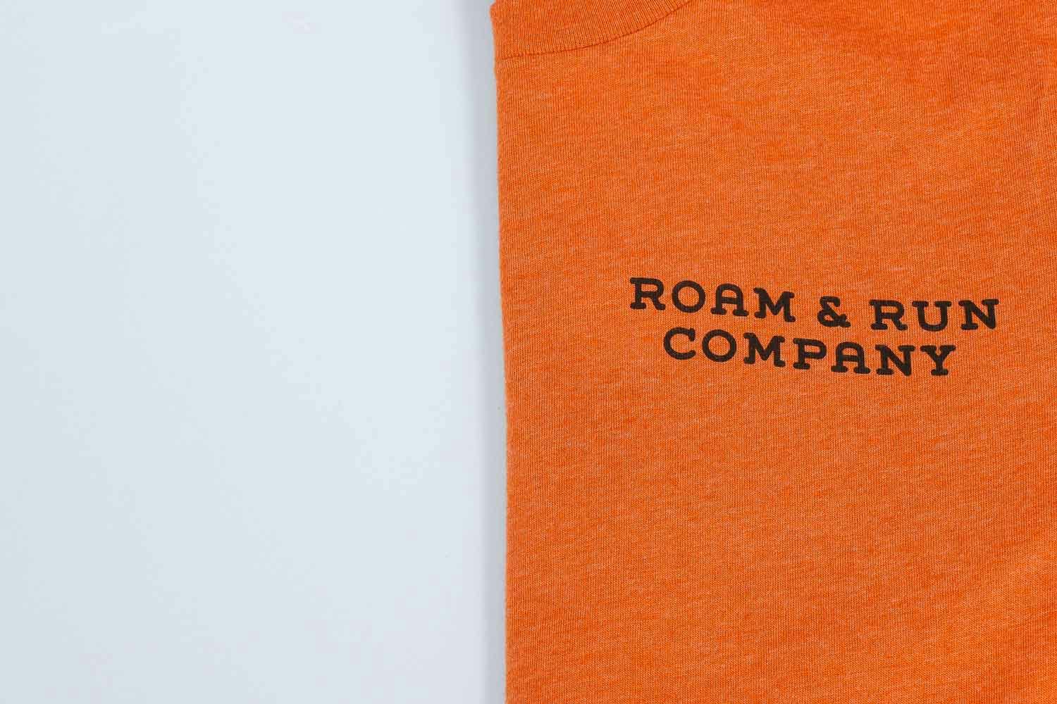 Orange shirt folded in half with "roam & run company" in black text on it Orange shirt folded in half with "roam & run company" in black text on it