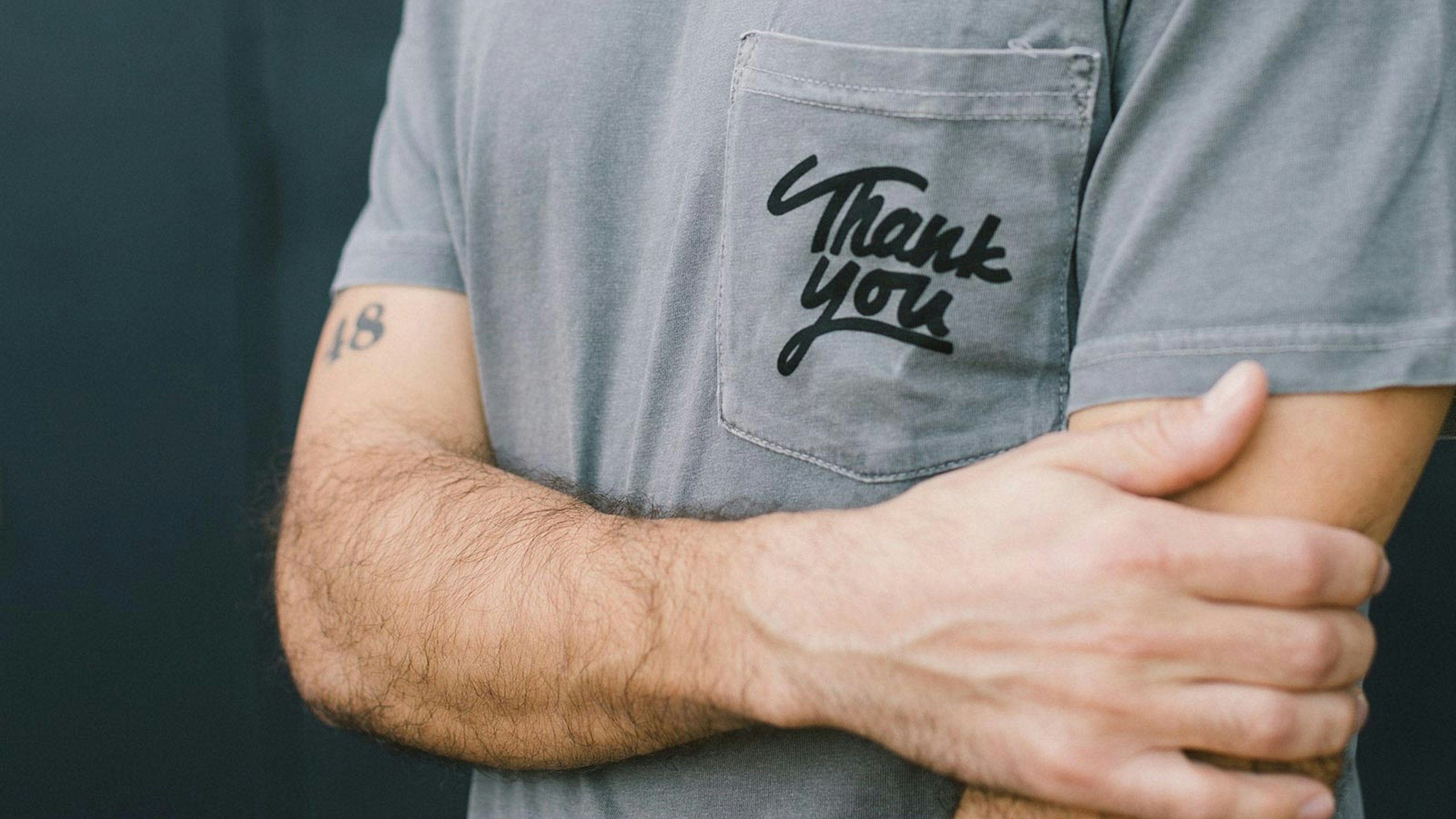 man cross arms wearing a gray shirt with the words thank you printed on the pocket