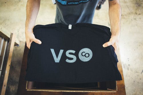 stack of black tees with white V S and logo being placed in a box