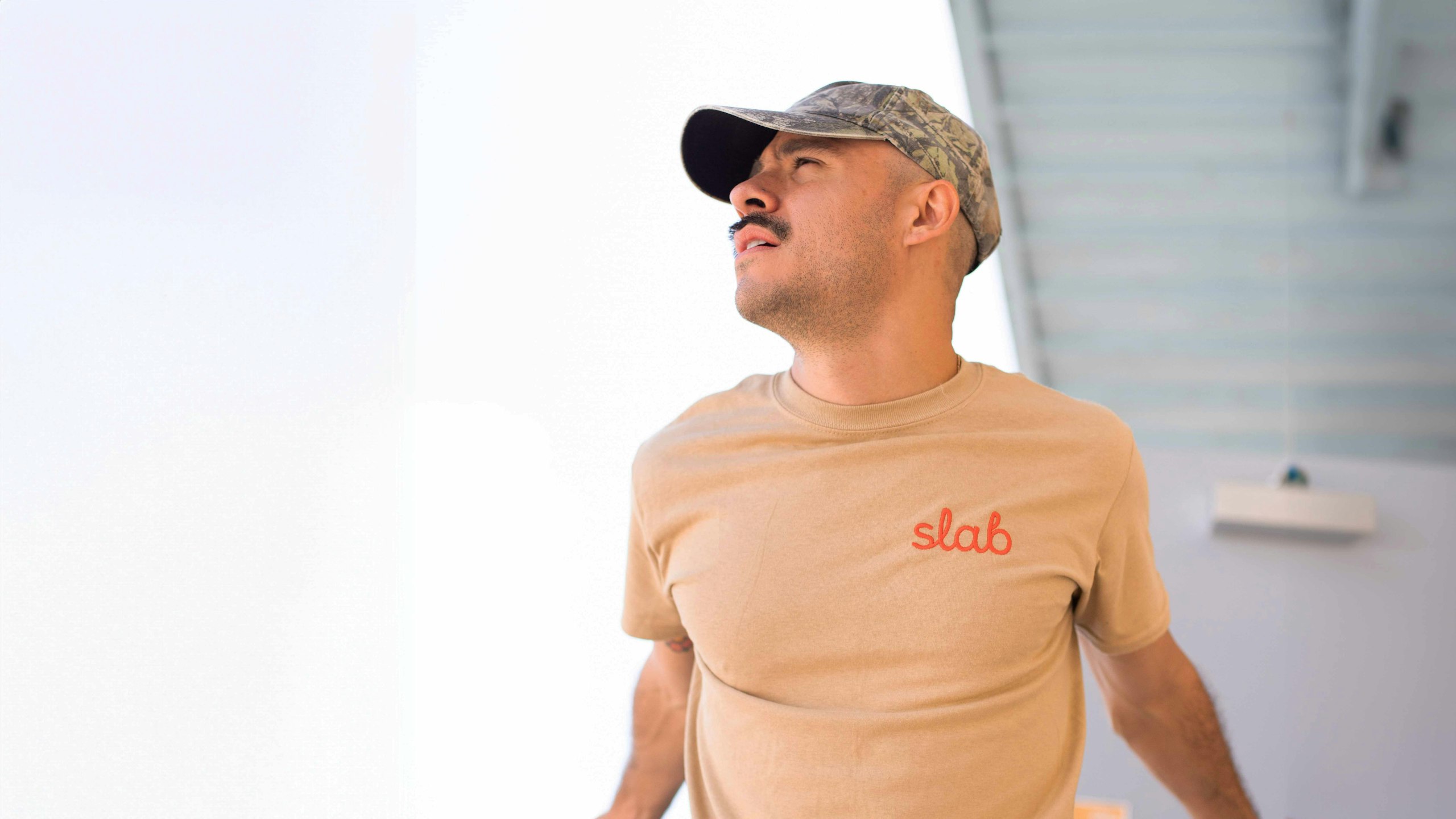 man wearing a tan t-shirt with slab embroidered on the top right in orange