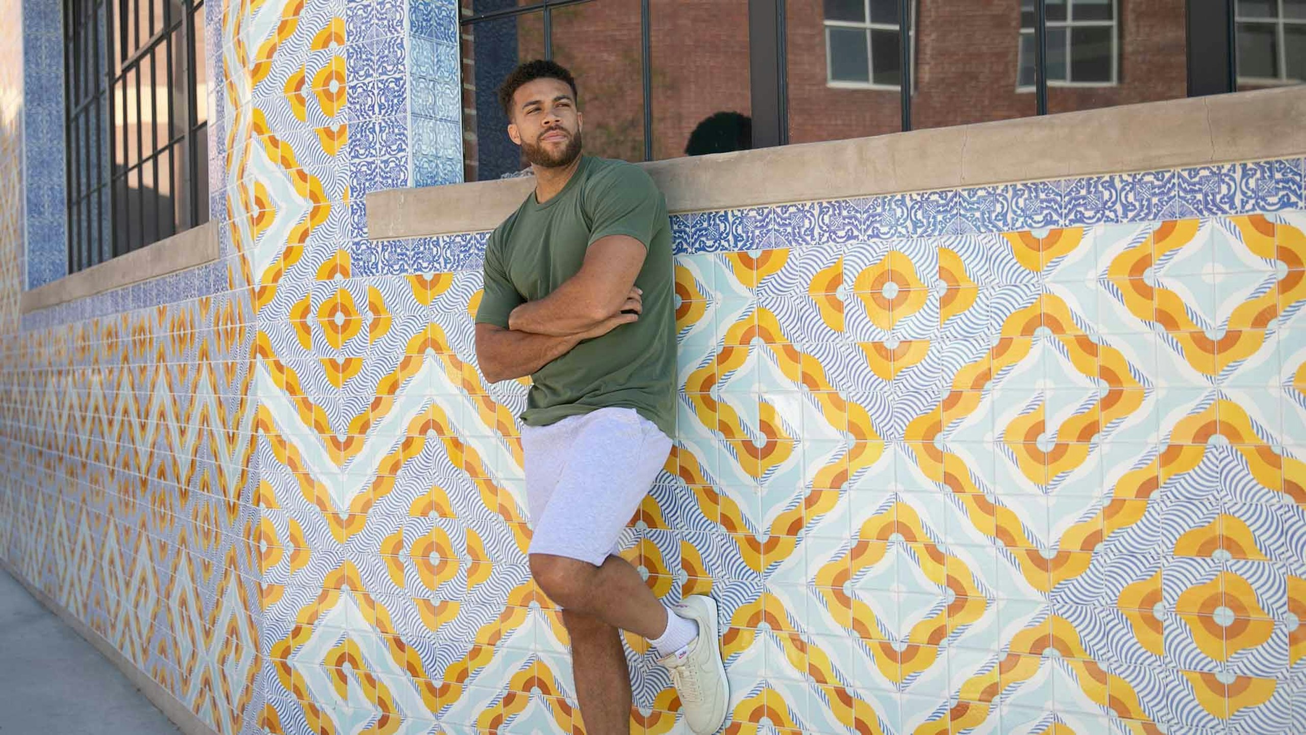 man wearing a dark green t-shirt and pink shorts leaning against a wall