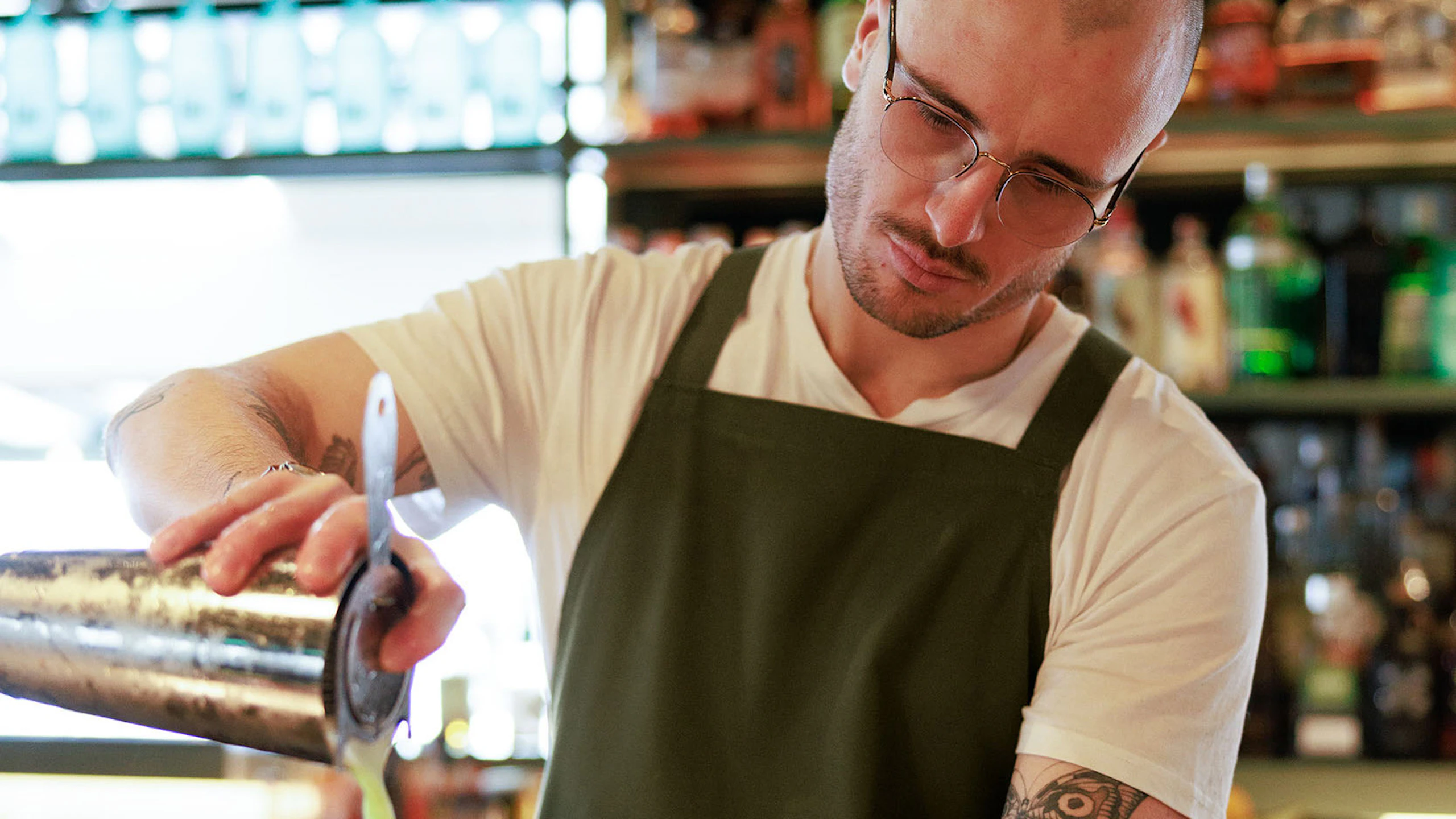 man wearing a white shirt with a olive green apron pouring a drink