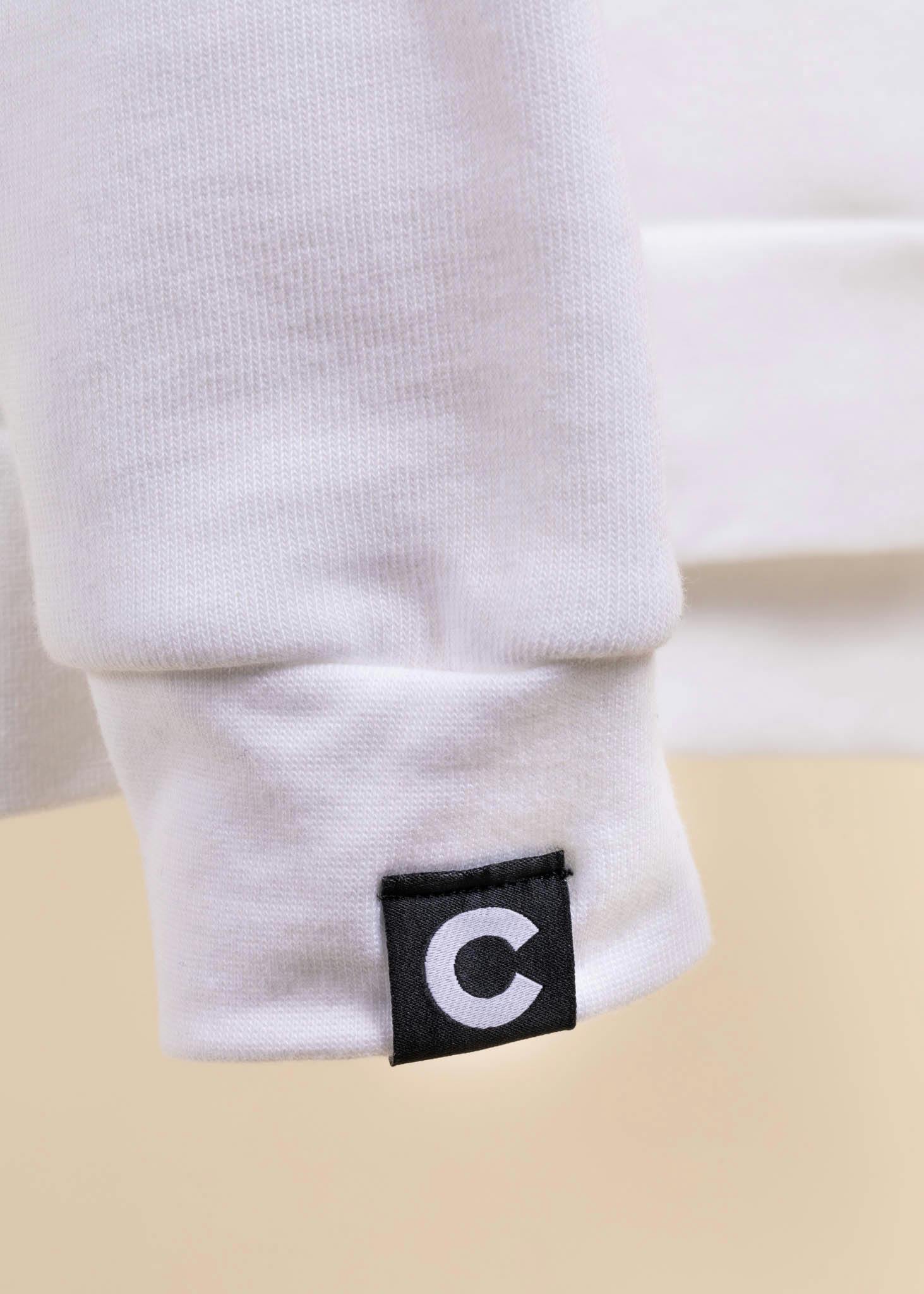 A black hem tag with a white "c" on it is attached to the sleeve of a white sweatshirt