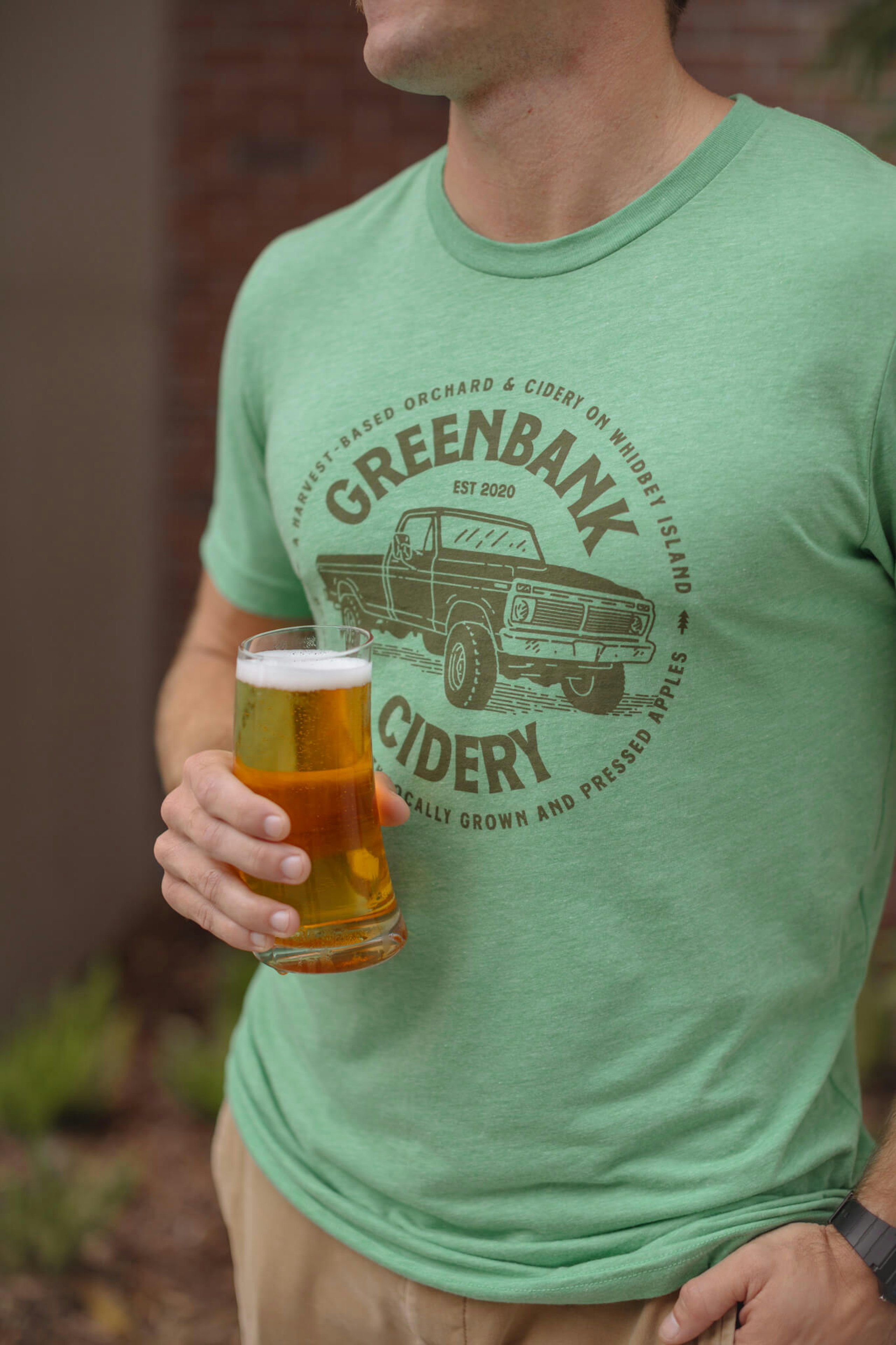 A man wears a green t-shirt for Greenbank Cidery while holding a glass of beer