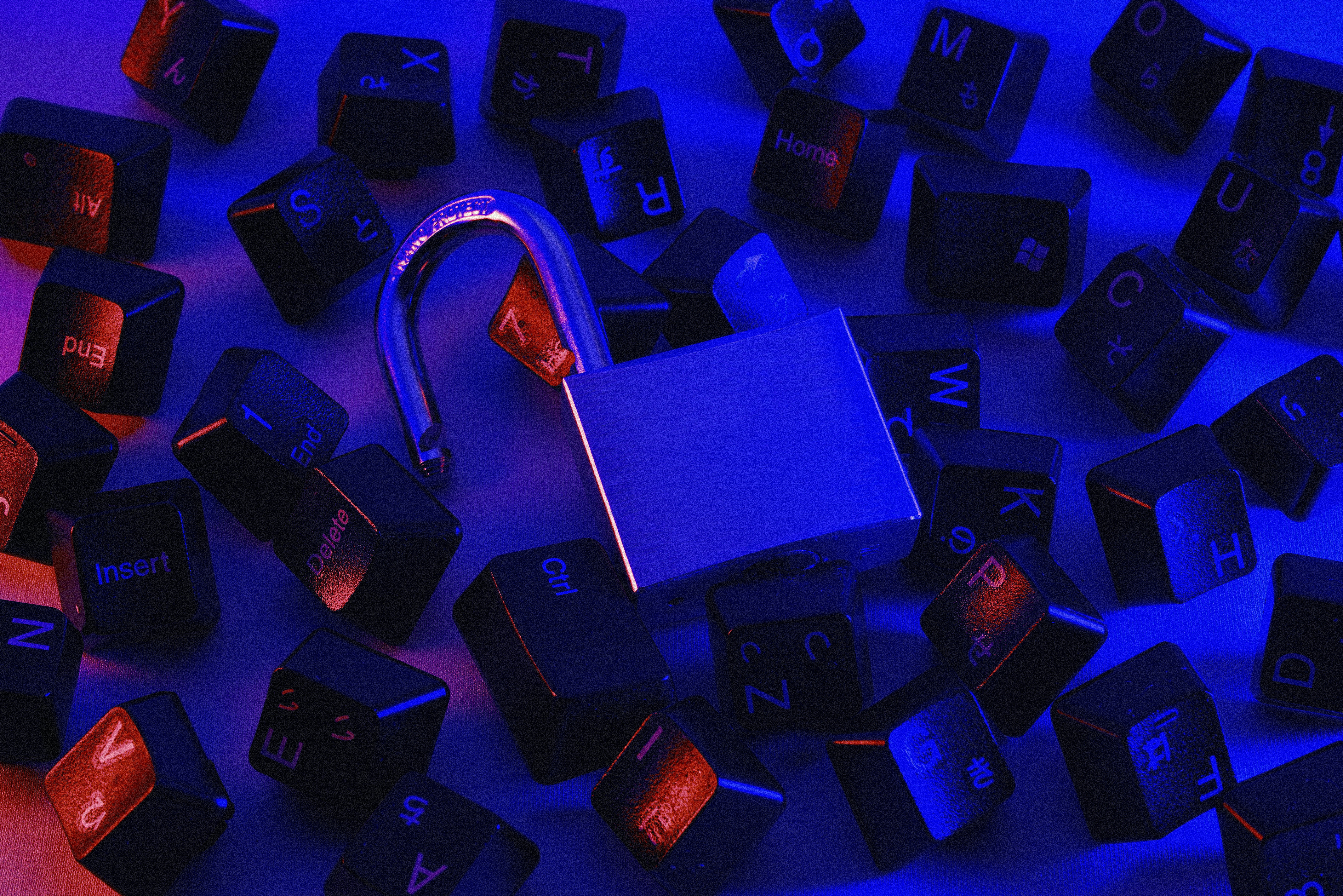 The image shows a close-up of a silver padlock that is slightly open, surrounded by scattered computer keyboard keys. The scene is bathed in a mix of blue and red lighting, creating a high-contrast, cyberpunk aesthetic. The keys feature various symbols and letters, hinting at a digital or cybersecurity theme, with the lock symbolizing protection or a breach in security. This visual is ideal for a website focusing on cybersecurity, fraud prevention, or digital security.